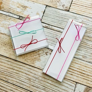May include: Two clear boxes with white cards inside. The boxes are tied with pink and teal ribbon bows. The text on the card reads "The Occasional Cookie".