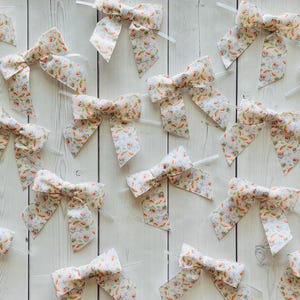 May include: A collection of fabric bows with a floral pattern featuring orange, green, and white elements. The bows are attached to clear plastic sticks, arranged on a white wooden surface.