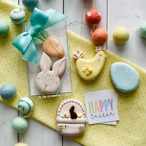 May include: A set of Easter-themed sugar cookies decorated with pastel colors. The cookies include a bunny, a chick, a basket with a bunny inside, and a carrot. The cookies are arranged on a yellow tablecloth with white polka dots. The text "Happy Easter" is written on a card in the image.