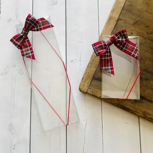 May include: Clear plastic gift boxes with red trim and plaid bows. The boxes are rectangular with a diagonal red line and are topped with a red, green, and gold plaid bow. One box is on a wooden surface, and the other is on a white wooden surface.