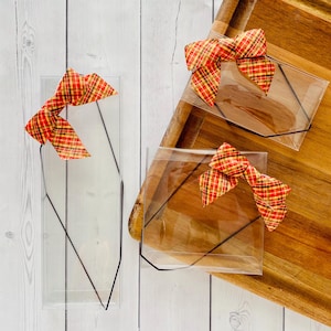 May include: Clear plastic packaging with a black outline and a plaid bow in red, yellow, and brown. The packaging is displayed on a wooden cutting board and a white wooden surface.