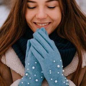 May include: Light blue gloves adorned with scattered white pearls. The gloves are made of a soft material and are worn by a person. The person is wearing a dark blue scarf and a brown coat with a white fluffy trim.
