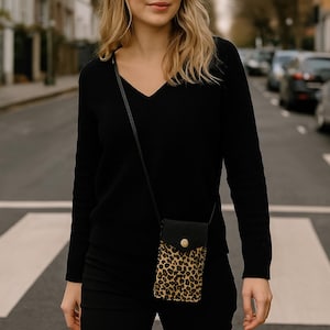 May include: A black crossbody phone bag with a leopard print design. The bag features a black flap with a gold button closure and a long black strap. The person is wearing a black v-neck sweater and black trousers.