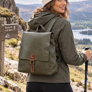 Puede incluir: Una mochila de color verde oliva con acabado texturizado, que presenta una solapa plegable asegurada por una correa de aspecto cuero marrón con una hebilla de metal color latón. La bolsa incluye un asa superior y correas ajustables para la espalda.