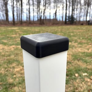 May include: A black square post cap sits atop a white square post. The cap has rounded edges and a textured surface. The background shows a blurred view of trees and grass.