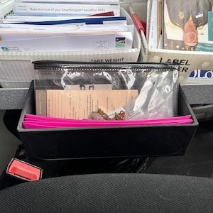 May include: A black plastic organizer with a clear zippered pouch and bright pink dividers. The organizer sits on a car seat, with various papers and documents visible in the background. A red seatbelt buckle is also visible.