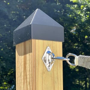 May include: A wooden post with a black cap and a stainless steel diamond-shaped eye plate. A metal ring is attached to the eye plate, connected to a gray fabric strap. The post is outdoors with a blurred green background.