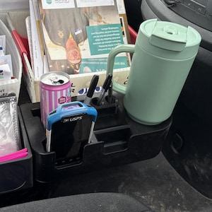 May include: A black car organizer with a mint green travel mug, a pink and white striped can, pens, and a USPS device. The organizer is filled with mail and other items.