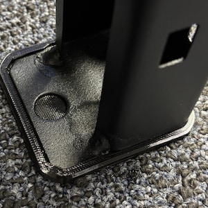 May include: Close-up of a black metal component with a textured base and a rectangular opening. The base has a circular indentation and a detailed border. The component is set against a gray and black patterned surface.