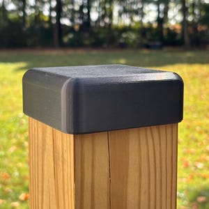 May include: A black, square post cap with rounded edges sits atop a wooden post. The post is made of light-colored wood with visible grain. The background is blurred, showing green grass and trees.