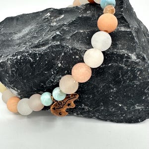 May include: A beaded bracelet featuring a variety of pastel-colored round beads, including peach, white, and light blue. A copper-colored charm in the shape of a frog is attached to the bracelet. The bracelet is displayed on a dark gray, textured stone.