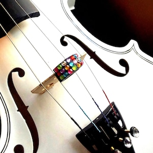 May include: Close-up of a white violin with black accents. The bridge is adorned with colorful, iridescent gems. The strings are visible, and the tuning pegs have clear, decorative ends. The instrument is set against a dark background.