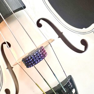 May include: Close-up of a white violin with black accents. The bridge is adorned with a decorative, jewel-encrusted piece in shades of purple and blue. The strings are visible, with a rainbow-colored string adding a pop of color. The instrument's f-holes are prominent.