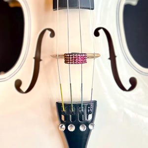 May include: Close-up of a white violin with a black fingerboard and bridge. The bridge is adorned with a decorative band of pink and gold rhinestones. The strings are visible, and the instrument has a decorative f-hole.