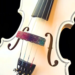 May include: A white violin with a black fingerboard and strings. The bridge is adorned with colorful rhinestones, transitioning from pink to blue. The instrument's body features classic f-holes and tuning pegs with crystal accents.