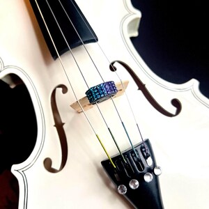 May include: A white violin with black accents and a decorative bridge. The bridge is adorned with a band of blue and purple rhinestones. The strings are visible, and the instrument is set against a dark background.