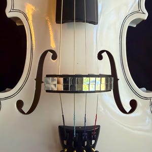 May include: Close-up of a white electric violin with black trim and a mirrored bridge. The strings are multi-colored, and the tuning pegs are clear, faceted crystals. The instrument has a glossy finish and is set against a dark background.