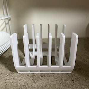 Peut inclure: Un égouttoir à vaisselle en plastique blanc avec des séparateurs verticaux. Le support a une forme rectangulaire avec des coins arrondis et est conçu pour maintenir les assiettes droites pour le séchage. Le support est posé sur un comptoir moucheté.