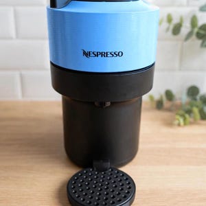 Könnte beinhalten: Eine Nespresso-Kaffeemaschine mit einem hellblauen oberen Gehäuse, auf dem das Nespresso-Logo in Schwarz aufgedruckt ist, einem soliden schwarzen Unterteil und einer runden schwarzen Abtropfschale mit perforierter Oberfläche an der Vorderseite.