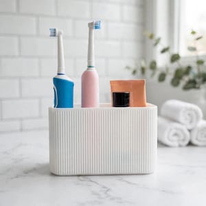 Puede incluir: Un organizador de baño rectangular blanco con textura acanalada vertical. El recipiente contiene dos cepillos de dientes eléctricos, uno con mango azul y otro con mango rosa, junto a un pequeño tubo color melocotón con tapa negra.