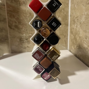 Könnte beinhalten: Ein weißer Lippenstift-Organizer aus Kunststoff, gefüllt mit verschiedenen Lippenstiften. Die Lippenstifte sind in verschiedenen Farben, darunter Rot, Braun und Schwarz. Einige Lippenstiftbehälter haben goldene oder schwarze Kappen. Der Organizer steht auf einer weißen Oberfläche.