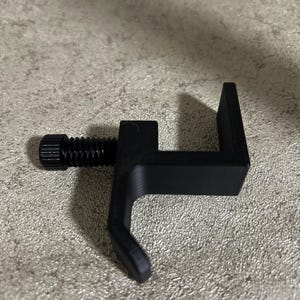 Puede incluir: Una abrazadera negra con un tornillo roscado y un diseño en forma de U. La abrazadera está hecha de un material sólido y está diseñada para asegurar objetos. El tornillo se utiliza para apretar la abrazadera. La abrazadera se muestra sobre una superficie texturizada.
