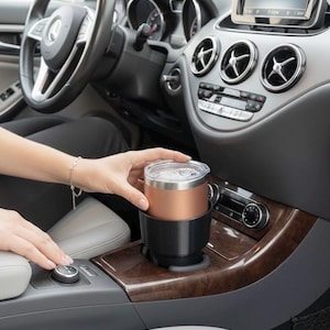 Könnte beinhalten: Ein kupferfarbener isolierter Reisebecher mit schwarzem Boden steht in einem Getränkehalter im Auto. Der Becher hat einen durchsichtigen Deckel und befindet sich in einem Autoinnenraum mit Holzverkleidung und grauen Akzenten. Das Armaturenbrett und das Lenkrad des Autos sind sichtbar.