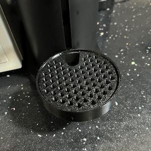 Puede incluir: Primer plano de una bandeja de goteo negra y redonda para cafetera. La bandeja tiene una superficie texturizada con numerosos agujeros pequeños. La bandeja está sobre una superficie negra moteada, con la base de la cafetera al fondo.