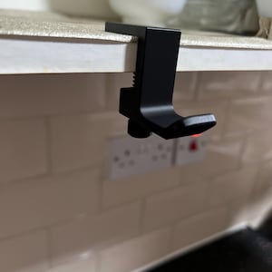 May include: A matte black metal under-shelf hook featuring an L-shaped design with a threaded screw mechanism for secure attachment to a shelf edge. The hook has a smooth finish and a sturdy construction, designed to hang items from the underside of a shelf.