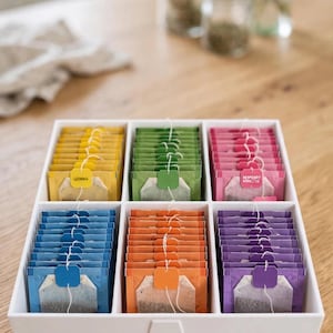 Op de afbeelding: Een witte rechthoekige theezakjeshouder met zes vakken, gevuld met theezakjes in verschillende kleuren: geel, groen, roze, blauw, oranje en paars. De houder is voorzien van een klein in reliëf aangebracht theekopje aan de voorzijde.