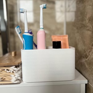 May include: A white, ribbed rectangular organiser on a white surface holds a blue and a pink electric toothbrush, a black jar, and a peach-coloured packet. A clear container with a wooden lid and cotton buds sits to the left.