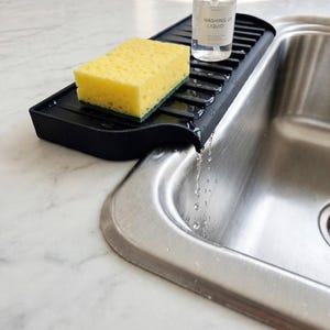 Puede incluir: Un organizador de fregadero de silicona negra con superficie acanalada y un pico de drenaje colocado sobre un fregadero de acero inoxidable. Una esponja rectangular amarilla descansa sobre el organizador junto a una botella.