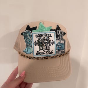 Puede incluir: Gorra de camionero beige con detalle de cadena dorada y parches bordados. Los parches incluyen una bota de vaquero turquesa, un frasco de perfume y un parche con el texto "Cowgirl Social Club". La gorra tiene una parte trasera de malla y una visera curva.
