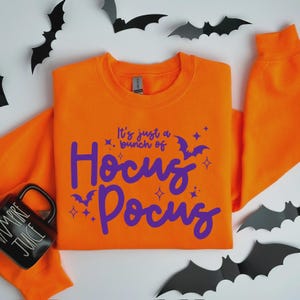 May include: An orange sweatshirt with the phrase "It's just a bunch of Hocus Pocus" in purple script. The design includes stars and bats. A black mug with "Vampire Juice" is on the left, and paper bats are scattered around.