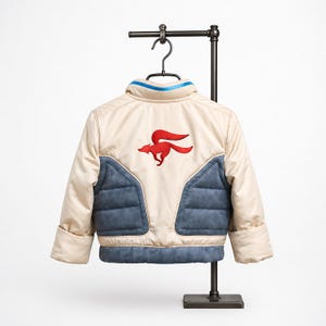 May include: Cream-colored children's jacket with a red fox logo on the back. The jacket features blue quilted panels and a blue stripe on the collar. The jacket is displayed on a black metal hanger.