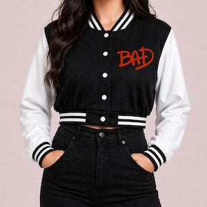 May include: Black and white varsity jacket with the word "BAD" in red script on the chest. The jacket has white sleeves, a black body, and white and black striped trim on the cuffs and waist. The jacket has white buttons.