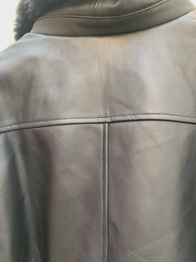 May include: Close-up of a black leather jacket's back, showcasing the collar, shoulder yoke, and vertical seam. The jacket features a shearling collar and detailed stitching, highlighting its craftsmanship. The leather appears smooth and supple.