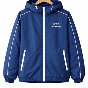 May include: A navy blue hooded jacket with white trim and a white zipper. The jacket has two front pockets and the words "KEEP SWIMMING" printed on the chest. The jacket is hanging on a wooden hanger.