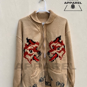 Op de afbeelding: Een beige gebreid vest met rits, geribbelde kraag en boorden. De voorzijde toont twee symmetrische rode en zwarte wolvenkopafbeeldingen, donkergrijze pootafdrukken bij de onderzoom en twee voorzakken. DARKLINE APPAREL logo.