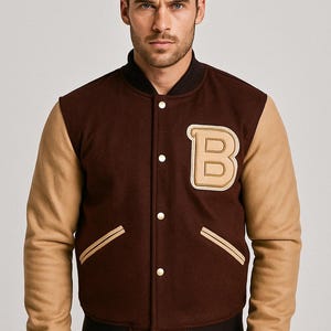 Puede incluir: Chaqueta universitaria marrón y beige con un parche grande con la letra "B". La chaqueta tiene un cuerpo marrón, mangas beige y puños y cuello de canalé negros. Los botones dorados y el ribete de los bolsillos añaden detalles.