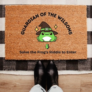 May include: A rectangular doormat made of natural coir fiber. The mat features a cartoon frog wearing a witch hat, with the text "GUARDIAN OF THE WILSONS" and "Solve the Frog's Riddle to Enter". Black boots are visible at the bottom.