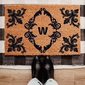May include: A rectangular doormat made of natural coir fiber. The mat features a black decorative design with the letter "W" in the center. The mat is placed on a black and white checkered rug, with a person's feet and black boots visible.