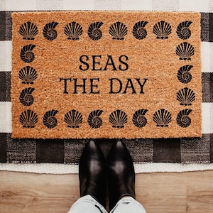May include: A rectangular doormat with the phrase "SEAS THE DAY" in black lettering, surrounded by a border of black seashell designs. The mat is made of a natural brown material and is placed on a black and white checkered rug. Black boots are visible.