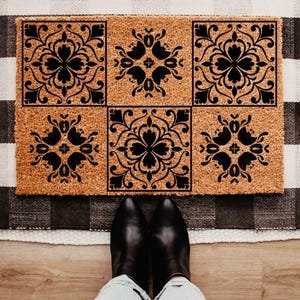 May include: A rectangular doormat with a natural coir fiber base features six black floral square designs. The mat is placed on a black and white checkered rug, with a person's feet in black boots visible at the bottom.