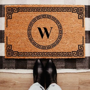 May include: A rectangular doormat with a Greek key border and a circular design in the center. The letter "W" is prominently displayed within the circle. The mat is brown with black detailing.