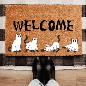 May include: A rectangular tan coir doormat featuring the black text WELCOME in a dripping font style. Below the text are four white cat illustrations, each wearing a white ghost costume with black eyes and black tails visible at the base.