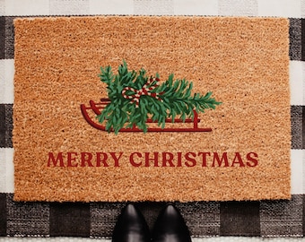 May include: A rectangular doormat with a brown coir surface. A red sled carries a green Christmas tree with a red and white candy cane bow. The words "MERRY CHRISTMAS" are in red. The mat is on a black and white checkered rug.