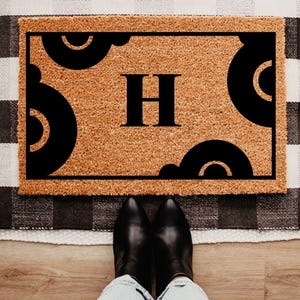 May include: A rectangular doormat with a natural coir surface features a black border and decorative black semi-circles. The letter "H" is prominently displayed in the center. Black boots and blue jeans are visible.