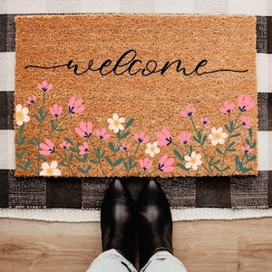 May include: A rectangular coir doormat with the word "welcome" in black script. The bottom edge features a floral design with pink and white flowers and green stems. Black boots and blue jeans are visible in the foreground.