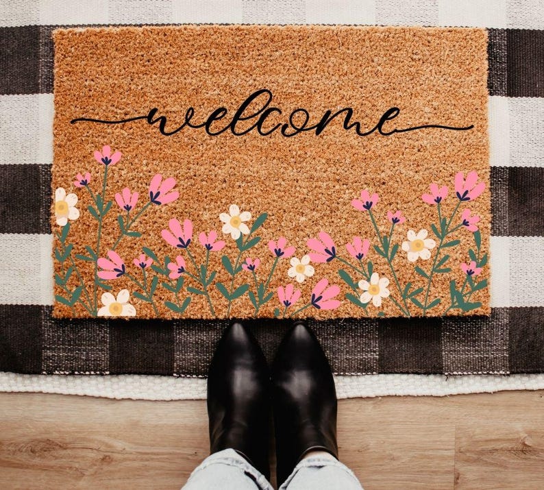 May include: A rectangular coir doormat with the word "welcome" in black script. The bottom edge features a floral design with pink and white flowers and green stems. Black boots and blue jeans are visible in the foreground.